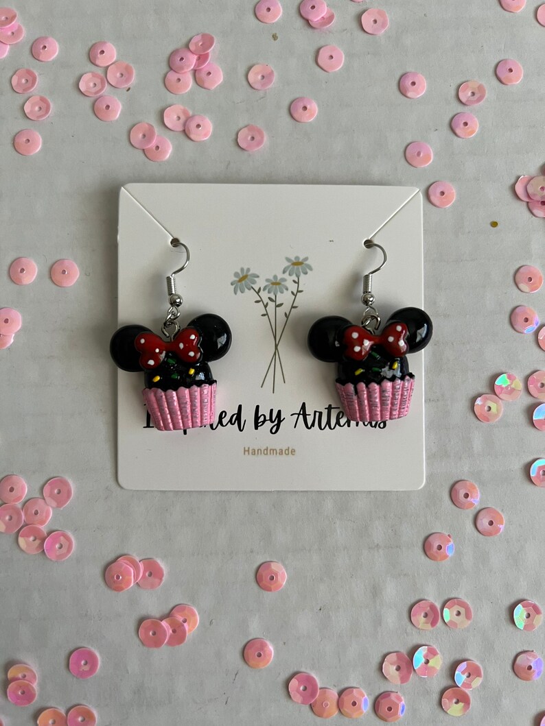 May include: A pair of black and pink cupcake earrings with a red bow and white polka dots. The earrings are handmade and feature a Mickey Mouse silhouette.