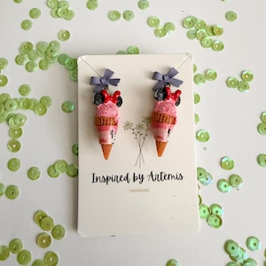 May include: A pair of handmade ice cream cone earrings with a pink scoop, a black and red bow, and a gray bow on top. The earrings are displayed on a white card with the text "Inspired by Artemis" and "Handmade".