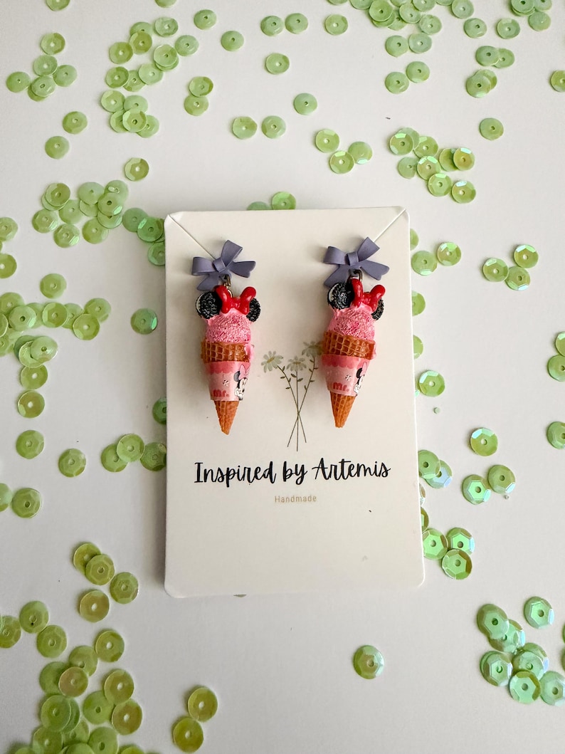 May include: A pair of handmade ice cream cone earrings with a pink scoop, a black and red bow, and a gray bow on top. The earrings are displayed on a white card with the text "Inspired by Artemis" and "Handmade".