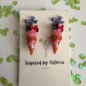 May include: A pair of novelty earrings shaped like pink ice cream cones with Minnie Mouse details. Each cone has a purple bow and a red bow. The earrings are displayed on a white card with the text "Inspired by Artemis Handmade".