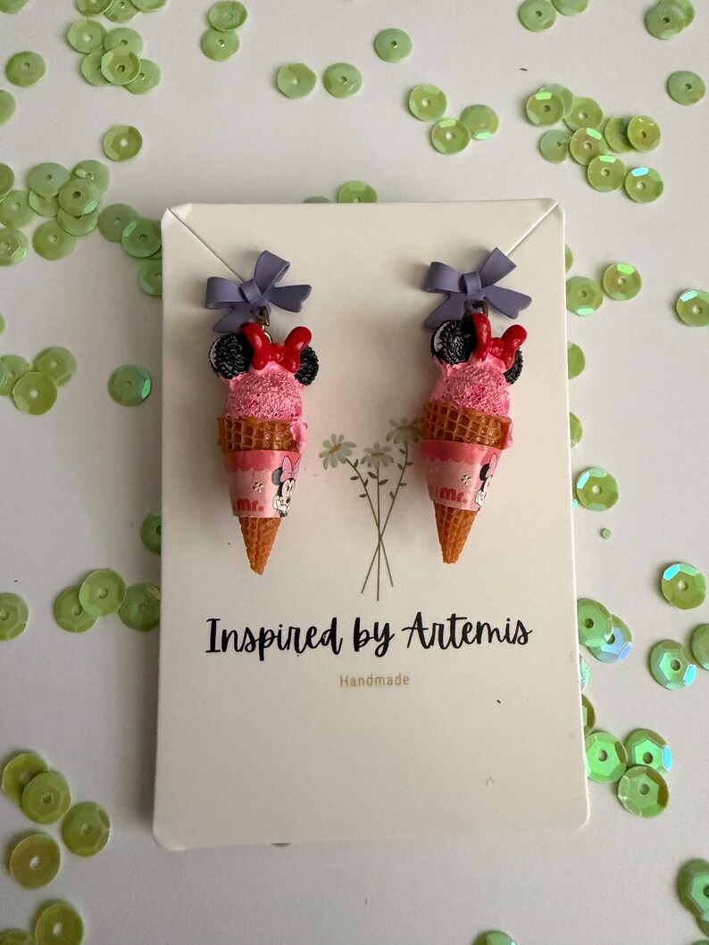 May include: A pair of novelty earrings shaped like pink ice cream cones with Minnie Mouse details. Each cone has a purple bow and a red bow. The earrings are displayed on a white card with the text "Inspired by Artemis Handmade".