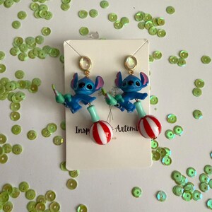 May include: A pair of blue Stitch earrings with red and white striped lollipop-shaped charms. The earrings have gold-colored ear wires and are displayed on a white card with the text "Inspired by Artemis". Green sequins are scattered around the earrings.