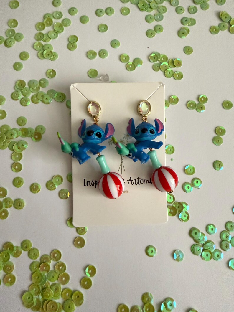 May include: A pair of blue Stitch earrings with red and white striped lollipop-shaped charms. The earrings have gold-colored ear wires and are displayed on a white card with the text "Inspired by Artemis". Green sequins are scattered around the earrings.