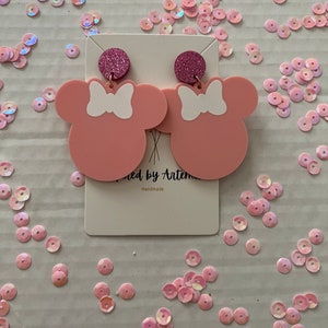 Puede incluir: Pendientes inspirados en Minnie Mouse de color rosa con lazos blancos y detalles de purpurina rosa. Los pendientes est&aacute;n hechos a mano y se muestran en una tarjeta blanca con el texto "Crafted by Artemis" y "Handmade".