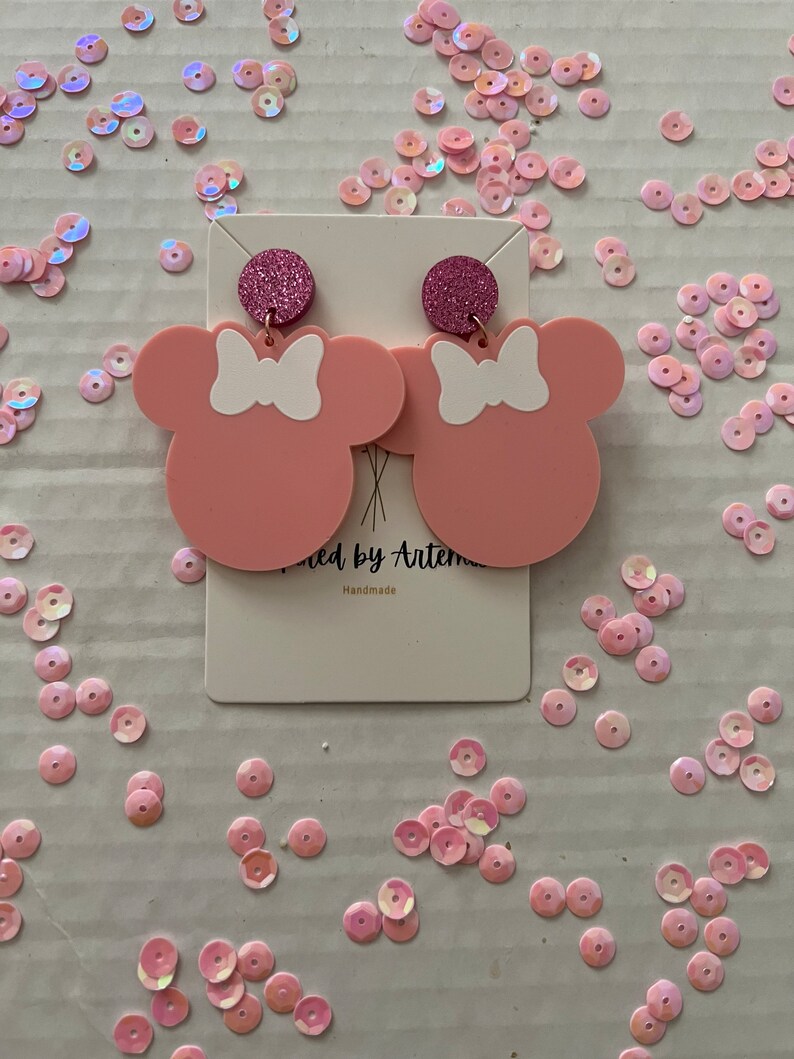 Puede incluir: Pendientes inspirados en Minnie Mouse de color rosa con lazos blancos y detalles de purpurina rosa. Los pendientes est&aacute;n hechos a mano y se muestran en una tarjeta blanca con el texto "Crafted by Artemis" y "Handmade".