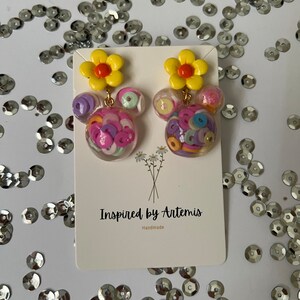 May include: A pair of handmade earrings featuring a yellow flower charm and a clear resin charm filled with colorful confetti. The earrings are displayed on a white card with the text "Inspired by Artemis Handmade".