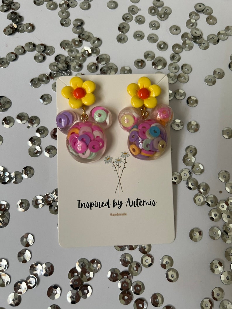 May include: A pair of handmade earrings featuring a yellow flower charm and a clear resin charm filled with colorful confetti. The earrings are displayed on a white card with the text "Inspired by Artemis Handmade".