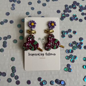 May include: Purple and green swirl dangle earrings with a gold bow and a purple flower charm. The earrings are on a white card with the text "Inspired by Artemis Handmade".