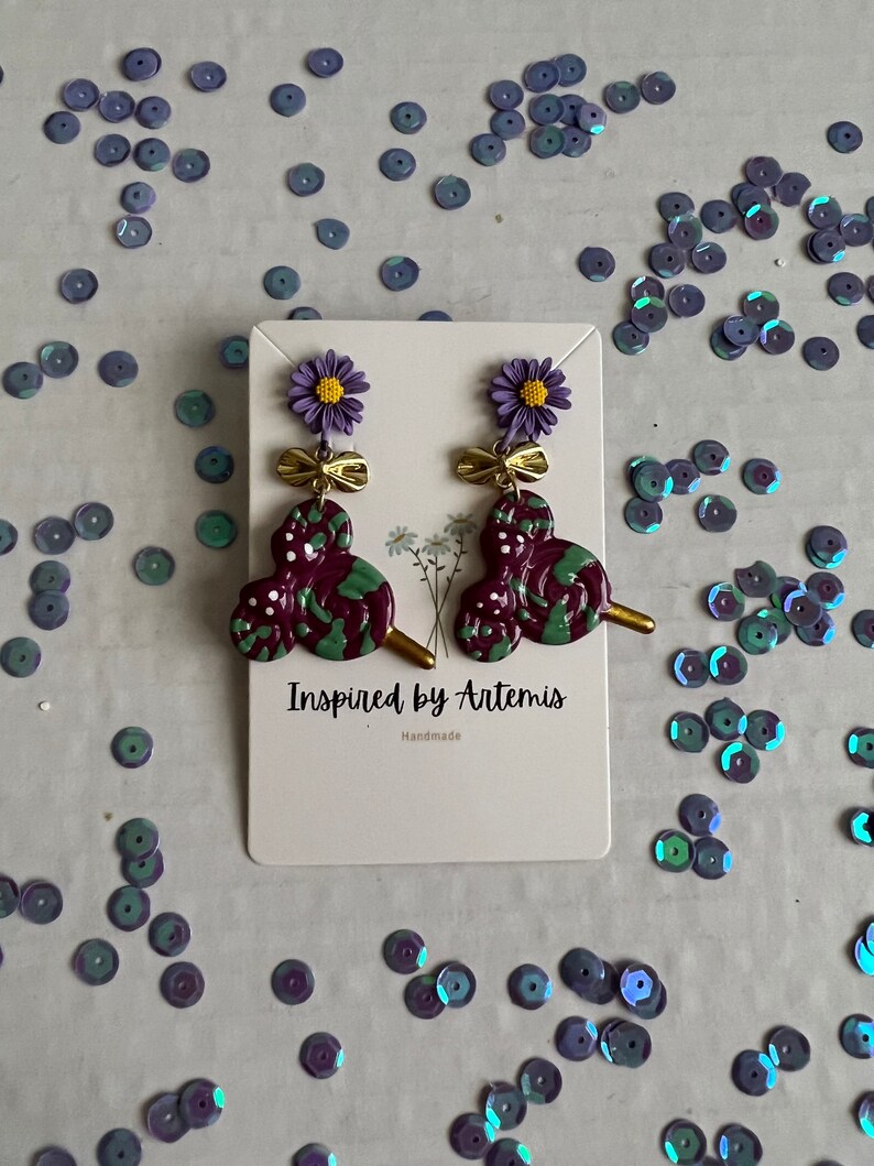 May include: Purple and green swirl dangle earrings with a gold bow and a purple flower charm. The earrings are on a white card with the text "Inspired by Artemis Handmade".