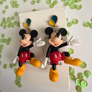 May include: A pair of Mickey Mouse earrings. Each earring features a black head with large ears, a red shorts, yellow shoes, and white gloves. The earrings are attached to a gold-colored hook with a blue and yellow accent.