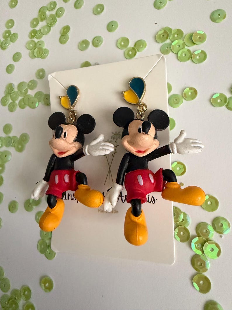 May include: A pair of Mickey Mouse earrings. Each earring features a black head with large ears, a red shorts, yellow shoes, and white gloves. The earrings are attached to a gold-colored hook with a blue and yellow accent.