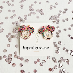 May include: A pair of gold-tone earrings featuring a cartoon character with a pink bow, black eyes, and a white face. The character is covered in colorful rhinestones. The earrings are on a white card with the text "Inspired by Artemis Handmade".