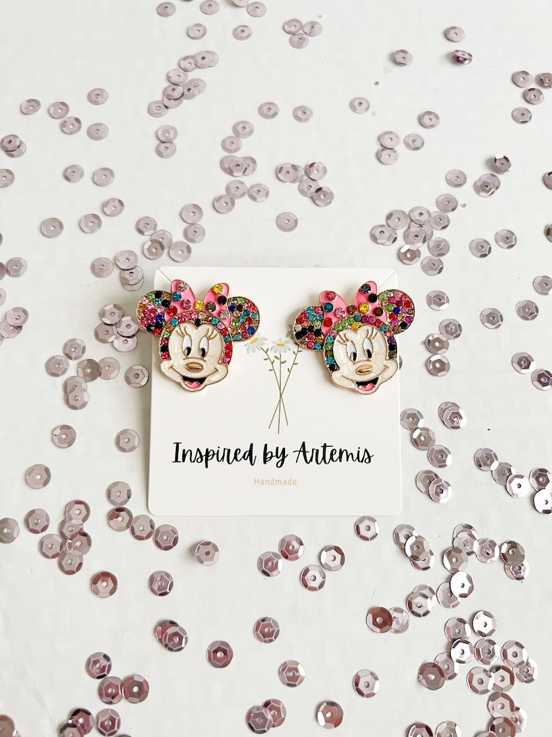 May include: A pair of gold-tone earrings featuring a cartoon character with a pink bow, black eyes, and a white face. The character is covered in colorful rhinestones. The earrings are on a white card with the text "Inspired by Artemis Handmade".