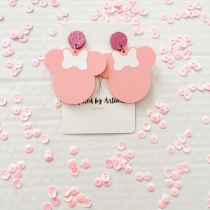 Puede incluir: Pendientes inspirados en Minnie Mouse de color rosa con lazos blancos y detalles de purpurina rosa. Los pendientes est&aacute;n en una tarjeta blanca con el texto "Wicked by Artemis Handmade".