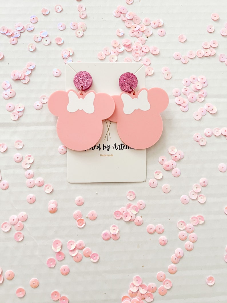 Puede incluir: Pendientes inspirados en Minnie Mouse de color rosa con lazos blancos y detalles de purpurina rosa. Los pendientes est&aacute;n en una tarjeta blanca con el texto "Wicked by Artemis Handmade".