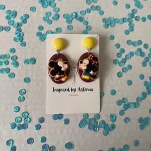 Puede incluir: Un par de pendientes hechos a mano con una imagen de dibujos animados de Mickey Mouse y Minnie Mouse. Los pendientes est&aacute;n hechos de arcilla polim&eacute;rica y tienen un fondo marr&oacute;n con detalles amarillos. El texto "Inspired by Artemis" y "Handmade" est&aacute; impreso en la tarjeta.