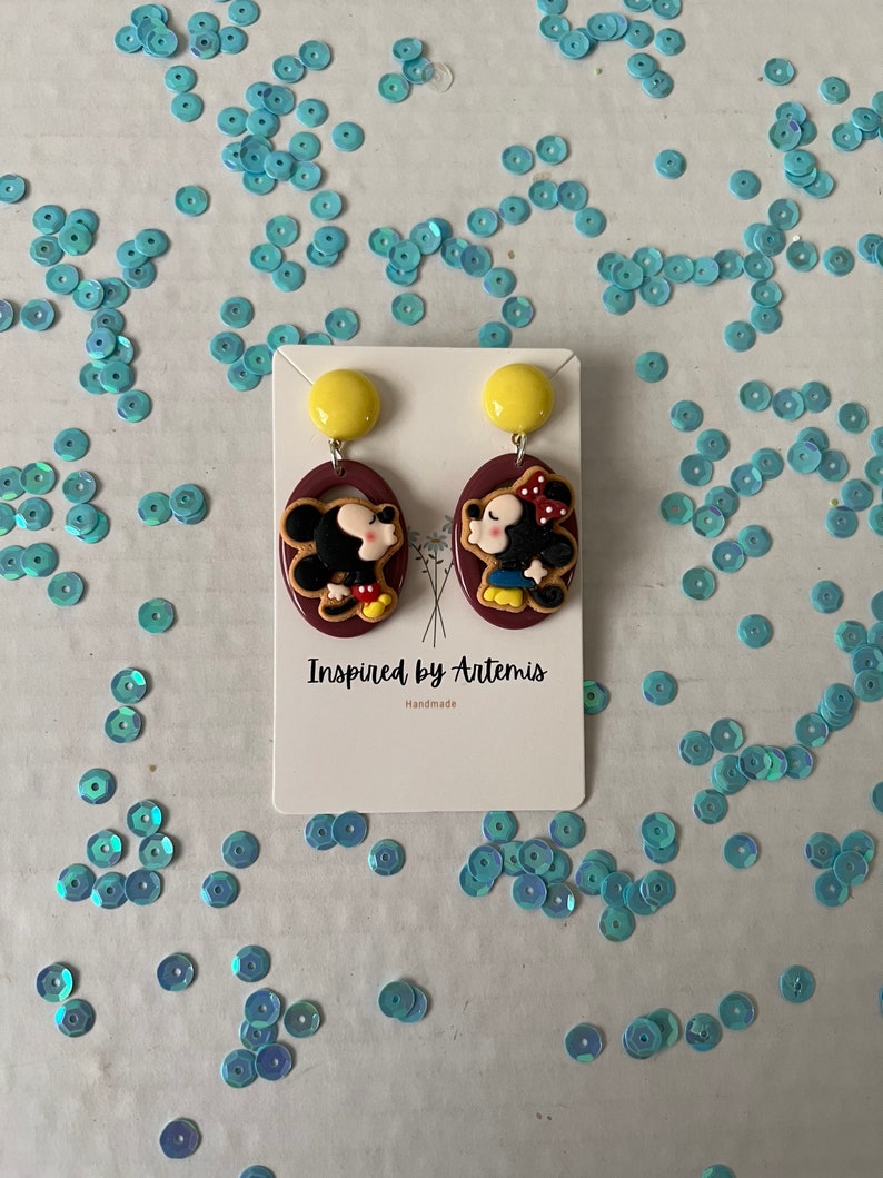 Puede incluir: Un par de pendientes hechos a mano con una imagen de dibujos animados de Mickey Mouse y Minnie Mouse. Los pendientes est&aacute;n hechos de arcilla polim&eacute;rica y tienen un fondo marr&oacute;n con detalles amarillos. El texto "Inspired by Artemis" y "Handmade" est&aacute; impreso en la tarjeta.