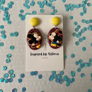 Puede incluir: Un par de pendientes hechos a mano con una imagen de dibujos animados de Mickey Mouse y Minnie Mouse bes&aacute;ndose. Los pendientes est&aacute;n hechos de arcilla polim&eacute;rica y tienen un fondo marr&oacute;n con detalles amarillos. Los pendientes est&aacute;n etiquetados como "Inspired by Artemis" y "Handmade".
