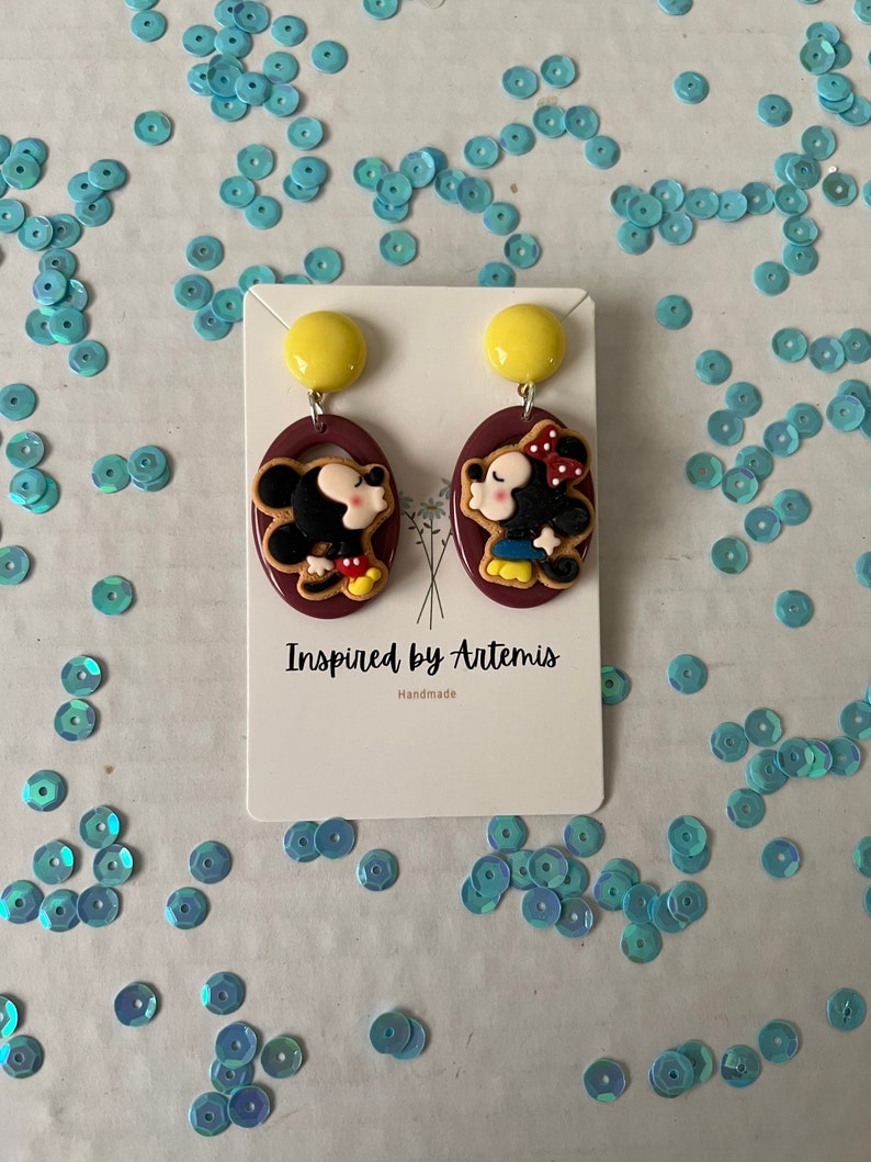 Puede incluir: Un par de pendientes hechos a mano con una imagen de dibujos animados de Mickey Mouse y Minnie Mouse bes&aacute;ndose. Los pendientes est&aacute;n hechos de arcilla polim&eacute;rica y tienen un fondo marr&oacute;n con detalles amarillos. Los pendientes est&aacute;n etiquetados como "Inspired by Artemis" y "Handmade".
