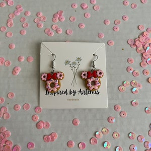 May include: A pair of donut-shaped earrings with pink frosting and colorful sprinkles. The earrings are shaped like Mickey Mouse ears and are handmade. The card reads "Inspired by Artemis Handmade".