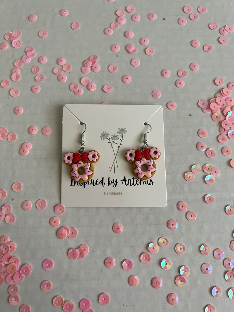 May include: A pair of donut-shaped earrings with pink frosting and colorful sprinkles. The earrings are shaped like Mickey Mouse ears and are handmade. The card reads "Inspired by Artemis Handmade".