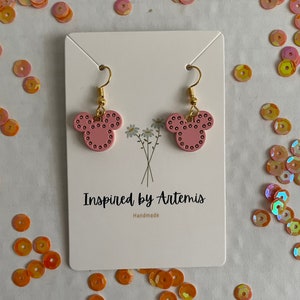 May include: Pink Mickey Mouse-shaped earrings with gold hooks and black dots. The earrings are displayed on a white card with the text "Inspired by Artemis" and "Handmade".