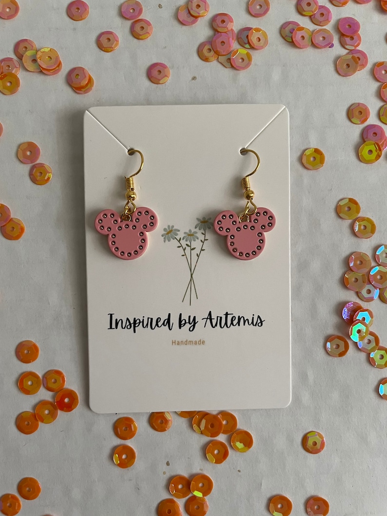 May include: Pink Mickey Mouse-shaped earrings with gold hooks and black dots. The earrings are displayed on a white card with the text "Inspired by Artemis" and "Handmade".