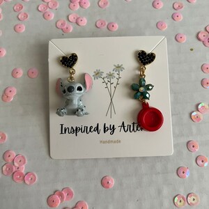 Peut inclure: Une paire de boucles d'oreilles faites &agrave; la main. Une boucle d'oreille pr&eacute;sente un clou en forme de c&oelig;ur noir, un personnage de dessin anim&eacute; gris et des accents dor&eacute;s. L'autre boucle d'oreille a un clou en forme de c&oelig;ur noir, une fleur verte et un charme circulaire rouge.