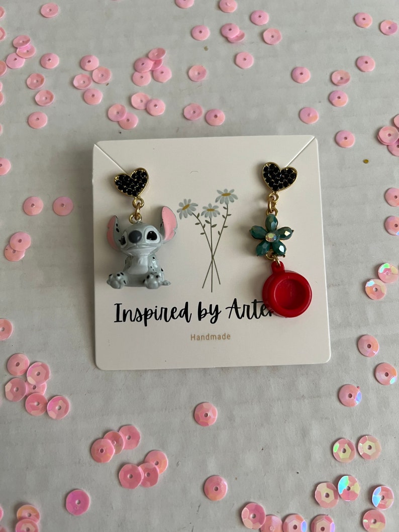 Peut inclure: Une paire de boucles d'oreilles faites &agrave; la main. Une boucle d'oreille pr&eacute;sente un clou en forme de c&oelig;ur noir, un personnage de dessin anim&eacute; gris et des accents dor&eacute;s. L'autre boucle d'oreille a un clou en forme de c&oelig;ur noir, une fleur verte et un charme circulaire rouge.