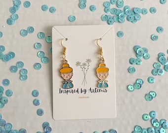 May include: A pair of gold dangle earrings featuring a cartoon character with a yellow hat, blue dress, and white collar. The earrings are on a white card with the text "Inspired by Artemis Handmade".
