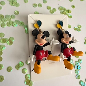 May include: A pair of Mickey Mouse earrings. The earrings feature a black head, red shorts, yellow shoes, and white gloves. Each earring has a gold-colored top with blue and yellow accents. The earrings are on a white card with the word "ine" and "Handmade" printed on it.