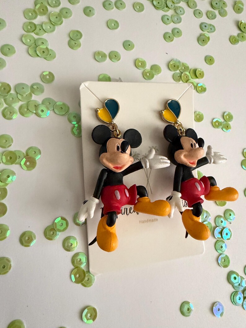 May include: A pair of Mickey Mouse earrings. The earrings feature a black head, red shorts, yellow shoes, and white gloves. Each earring has a gold-colored top with blue and yellow accents. The earrings are on a white card with the word "ine" and "Handmade" printed on it.