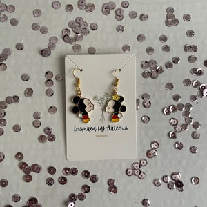 May include: Gold-tone earrings featuring a cartoon couple kissing. The earrings are on a white card with the text "Inspired by Artemis" and "Handmade".