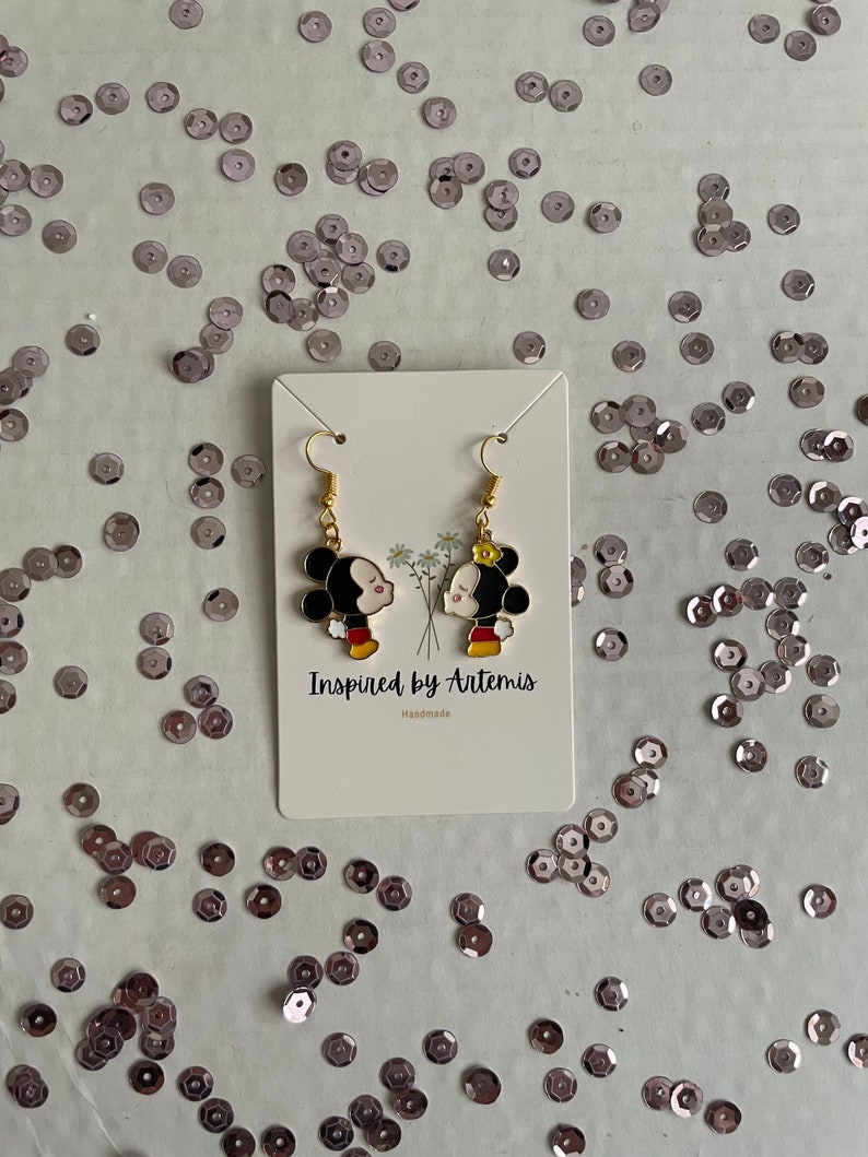 May include: Gold-tone earrings featuring a cartoon couple kissing. The earrings are on a white card with the text "Inspired by Artemis" and "Handmade".