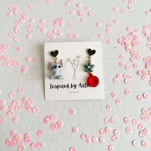 Peut inclure: Boucles d'oreilles faites &agrave; la main avec un personnage de dessin anim&eacute; et des breloques. Une boucle d'oreille repr&eacute;sente un personnage de dessin anim&eacute; gris et blanc, l'autre une fleur verte et un charme rouge. Les boucles d'oreilles sont sur une carte blanche avec le texte "Inspired by Arte".