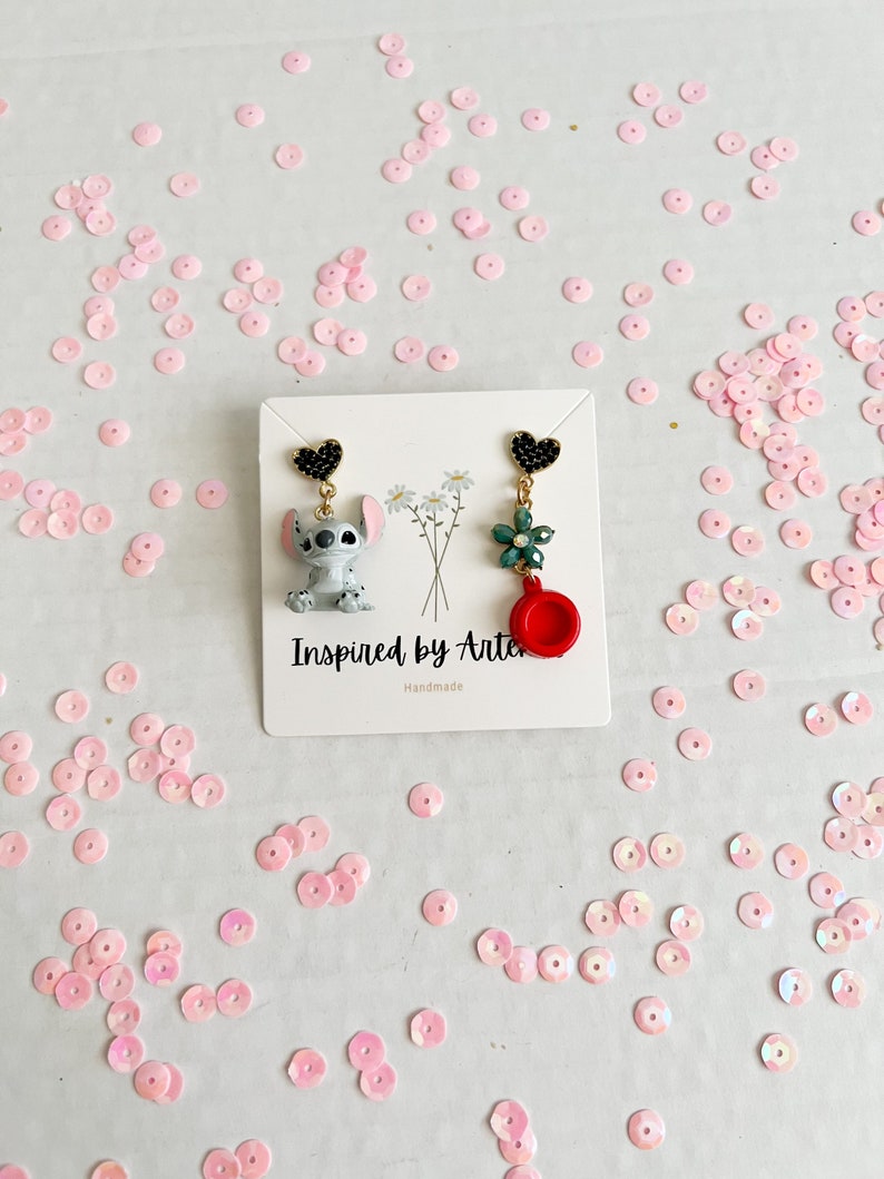 Peut inclure: Boucles d'oreilles faites &agrave; la main avec un personnage de dessin anim&eacute; et des breloques. Une boucle d'oreille repr&eacute;sente un personnage de dessin anim&eacute; gris et blanc, l'autre une fleur verte et un charme rouge. Les boucles d'oreilles sont sur une carte blanche avec le texte "Inspired by Arte".