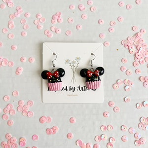 May include: A pair of black and red cupcake earrings with white frosting and a red bow. The earrings are on a white card with the text "Inspired by Artemis" and "Handmade".