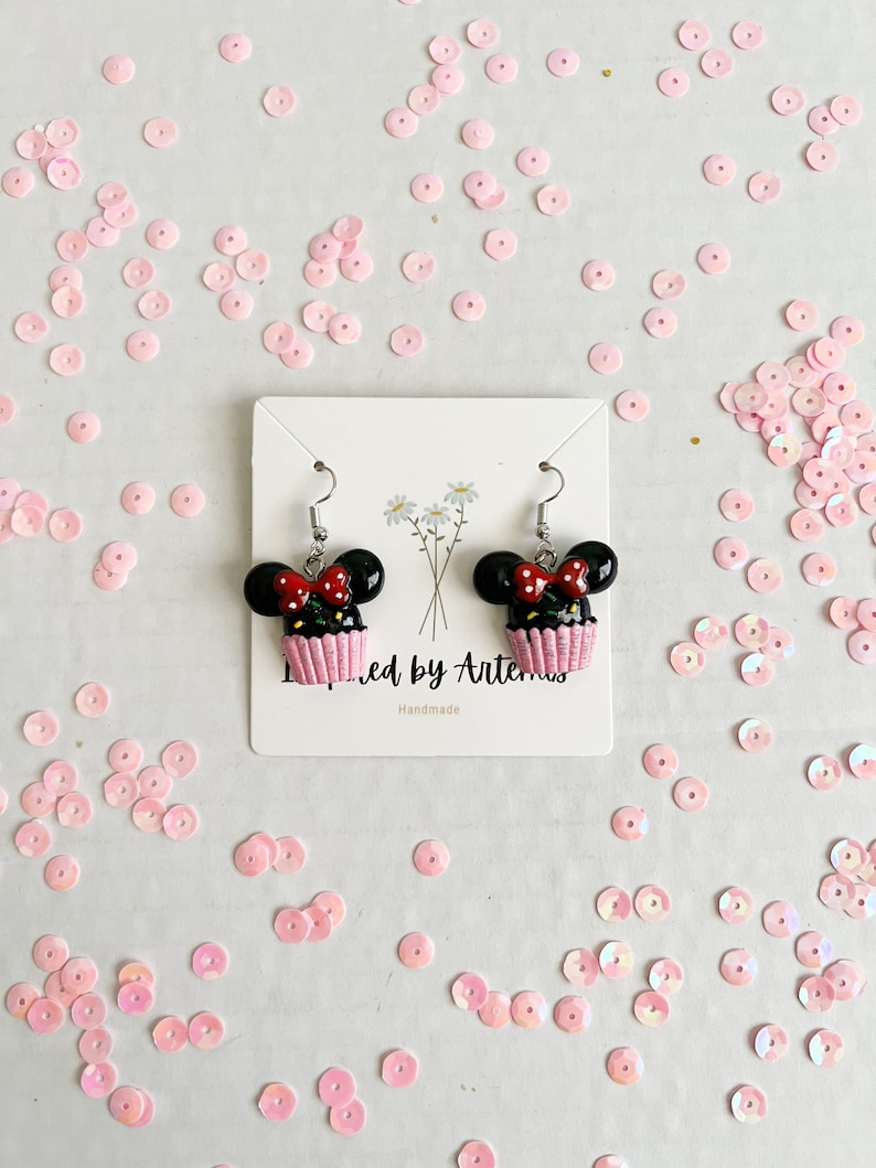 May include: A pair of black and red cupcake earrings with white frosting and a red bow. The earrings are on a white card with the text "Inspired by Artemis" and "Handmade".