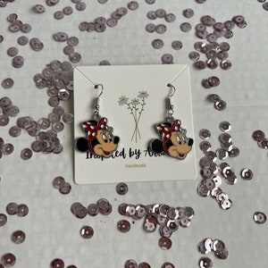 May include: A pair of silver dangle earrings featuring a cartoon image of Minnie Mouse with red and white polka dots. The earrings are on a white card with the text "Inspired by Ava" and "Handmade".