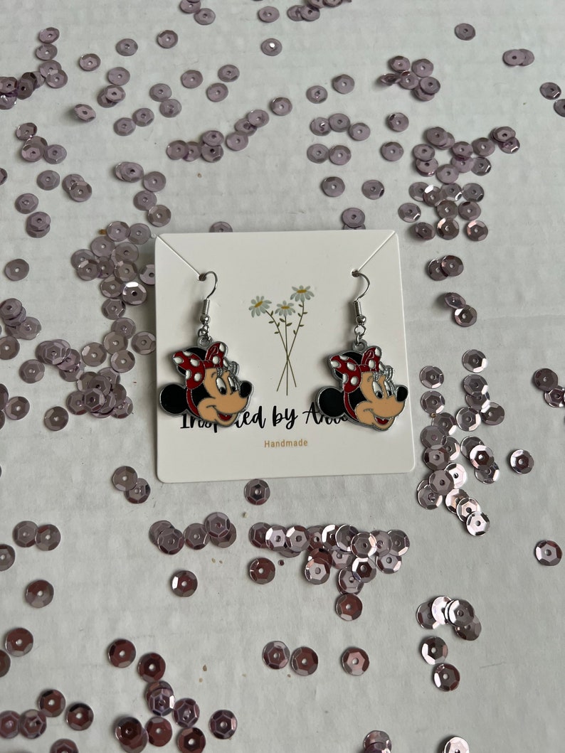 May include: A pair of silver dangle earrings featuring a cartoon image of Minnie Mouse with red and white polka dots. The earrings are on a white card with the text "Inspired by Ava" and "Handmade".