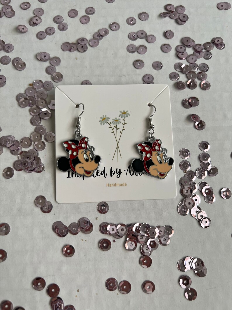 May include: A pair of silver earrings with red and white Minnie Mouse heads. The earrings are on a white card with the text "Inspired by Ava" and "Handmade".