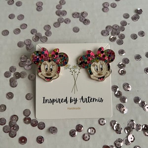May include: A pair of gold-tone earrings featuring a colorful, jeweled Minnie Mouse design. The earrings are on a white card with the text "Inspired by Artemis" and "Handmade".