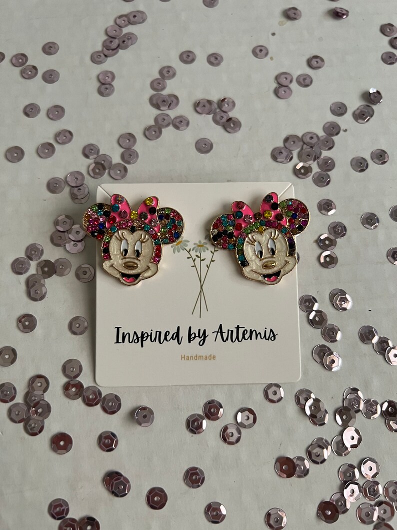 May include: A pair of gold-tone earrings featuring a colorful, jeweled Minnie Mouse design. The earrings are on a white card with the text "Inspired by Artemis" and "Handmade".