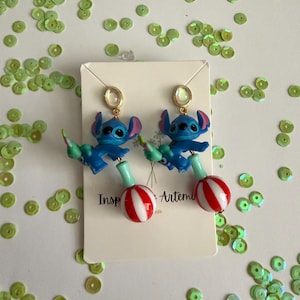 May include: A pair of blue Stitch earrings with gold-colored studs. Each earring features a Stitch figure holding a green and red striped lollipop. The earrings are on a white card with the text "Inspired by Artemis".