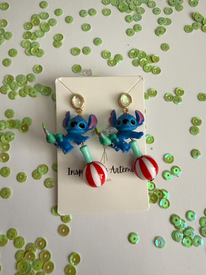 May include: A pair of blue Stitch earrings with gold-colored studs. Each earring features a Stitch figure holding a green and red striped lollipop. The earrings are on a white card with the text "Inspired by Artemis".