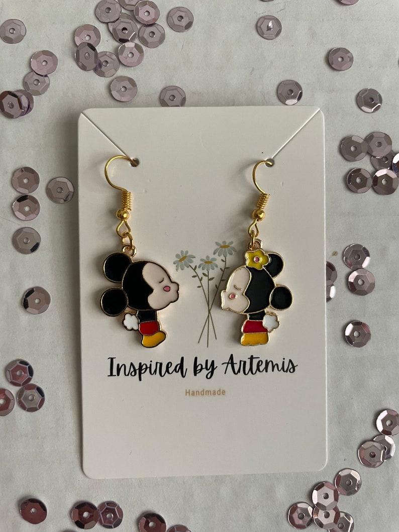 May include: A pair of gold dangle earrings featuring a cartoon couple kissing. The characters are Mickey Mouse and Minnie Mouse. The earrings are handmade and the card reads "Inspired by Artemis".
