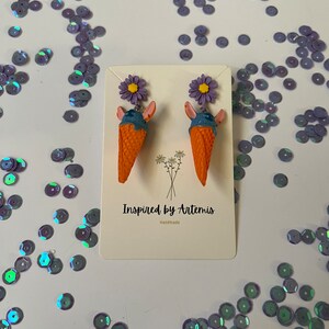 May include: A pair of ice cream cone earrings with purple flowers. The earrings are handmade and are made of polymer clay. The ice cream cones are orange and the ice cream is blue. The flowers are purple and have yellow centers. The earrings are on a white background with the text "Inspired by Artemis Handmade".