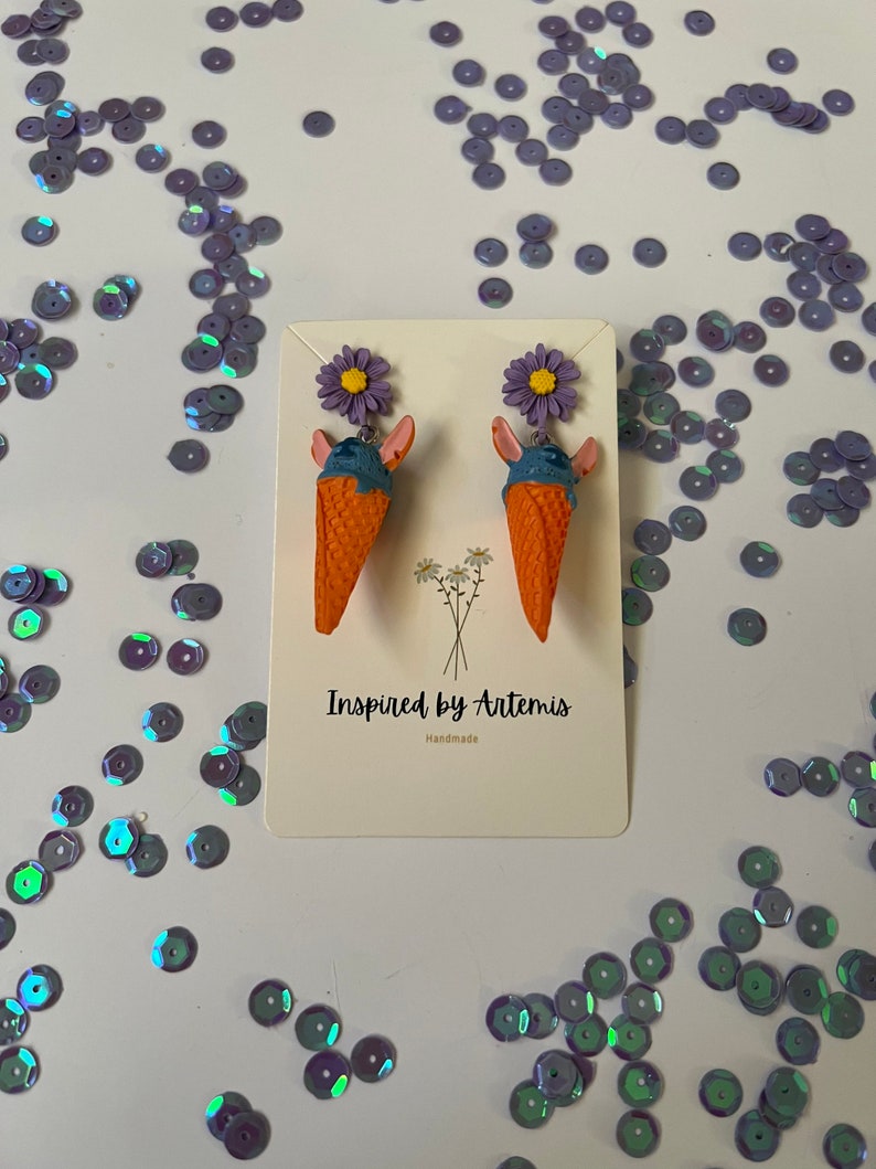 May include: A pair of ice cream cone earrings with purple flowers. The earrings are handmade and are made of polymer clay. The ice cream cones are orange and the ice cream is blue. The flowers are purple and have yellow centers. The earrings are on a white background with the text "Inspired by Artemis Handmade".