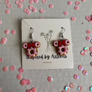May include: A pair of handmade donut earrings with pink frosting, sprinkles, and red bows. The earrings are on a white card with the text "Inspired by Artemis" and "Handmade".