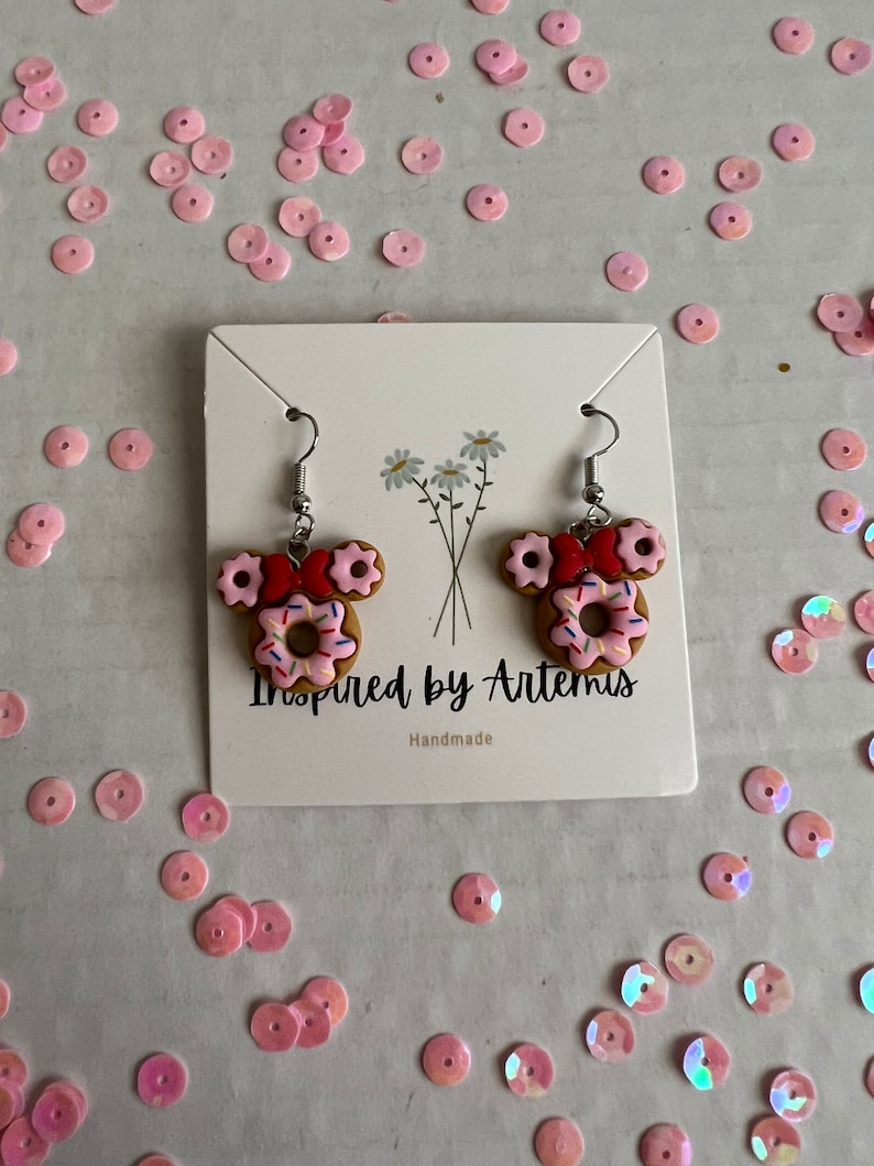 May include: A pair of handmade donut earrings with pink frosting, sprinkles, and red bows. The earrings are on a white card with the text "Inspired by Artemis" and "Handmade".